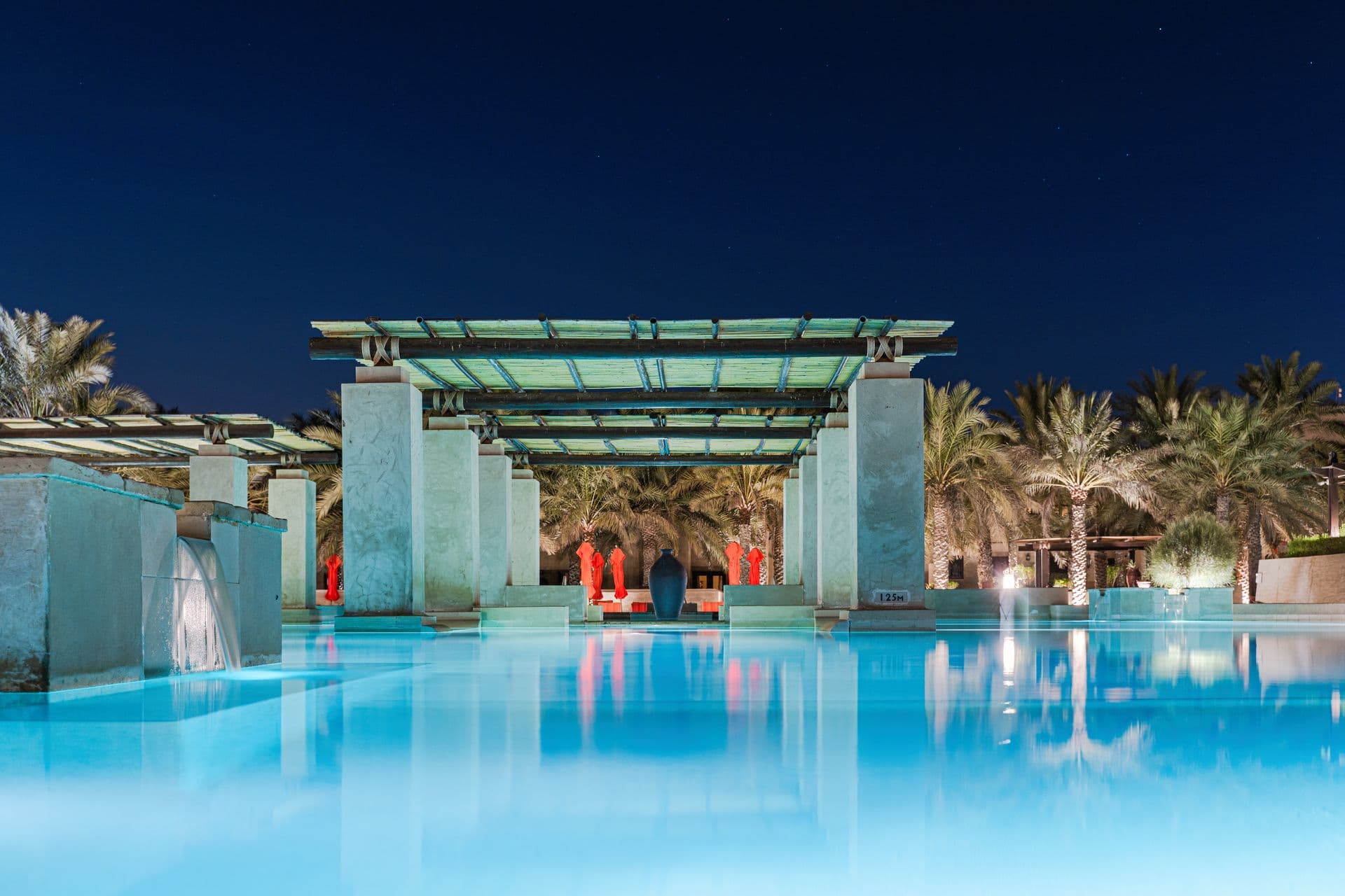 The resort pool lit at night, palms and pergola beyond